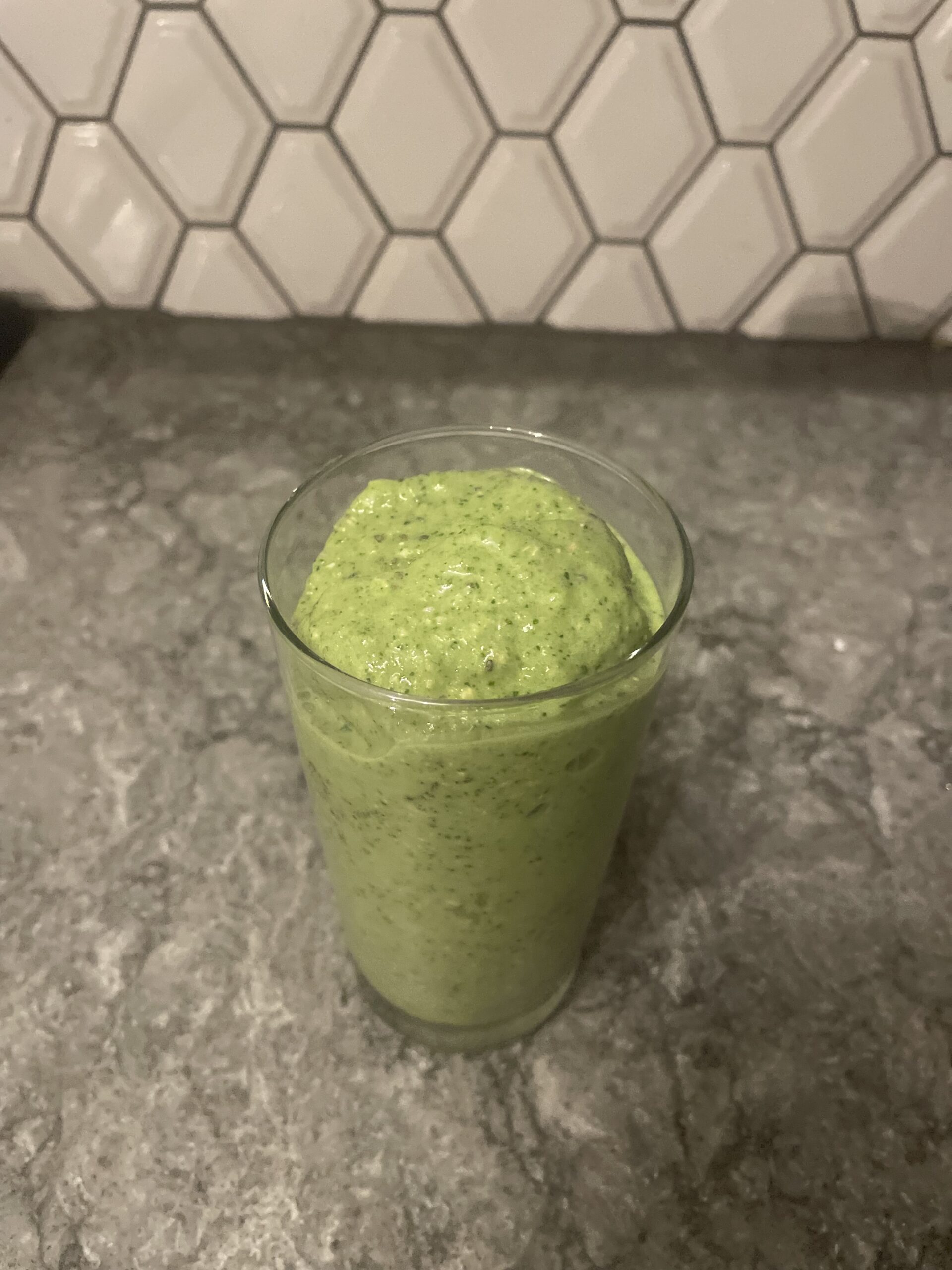 Green smoothie in glass on gray kitchen counter with white hexagonal backsplash.